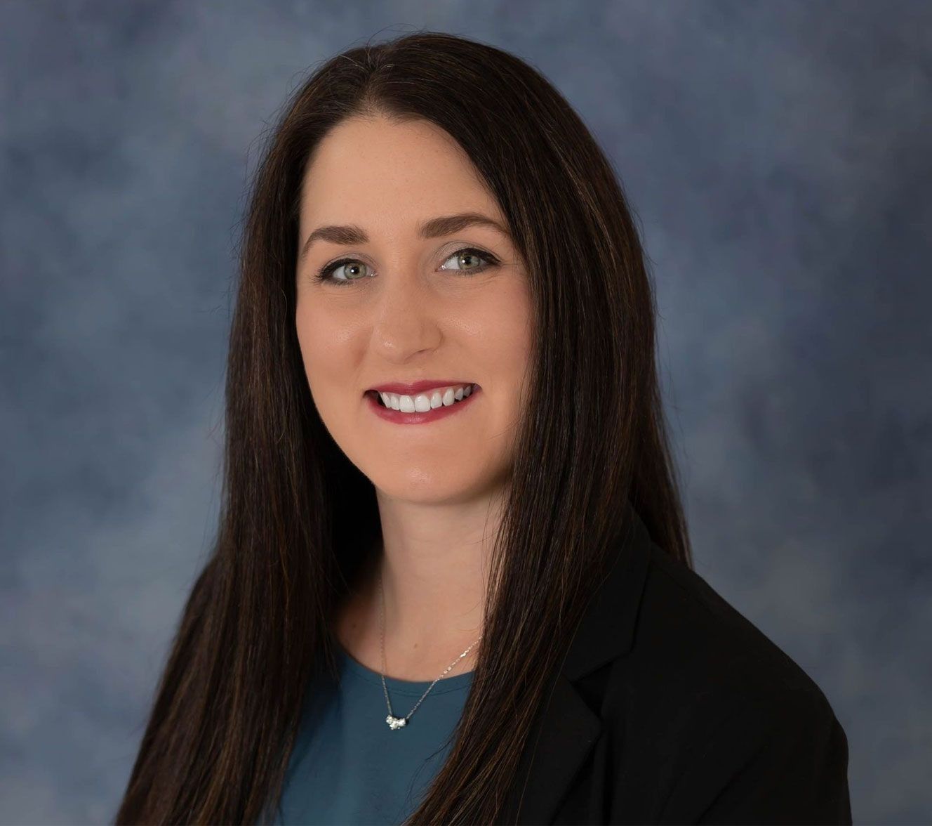 Professional headshot of a smiling person with long dark hair, wearing a teal top and black blazer against a blue backdrop.