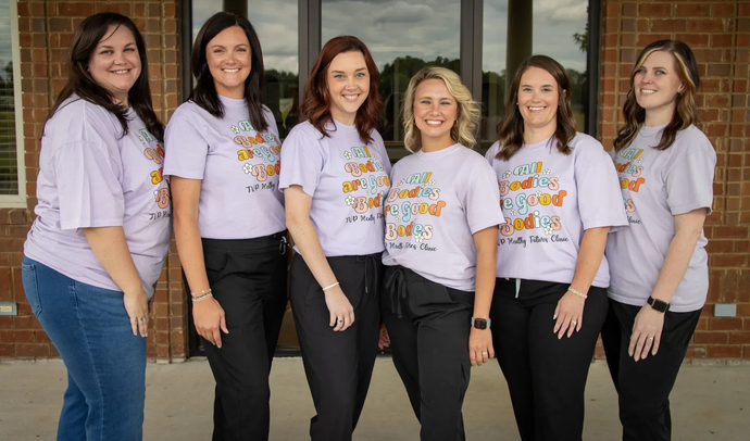 Six people standing in a row in front of a brick building, wearing matching light purple t-shirts and dark pants.