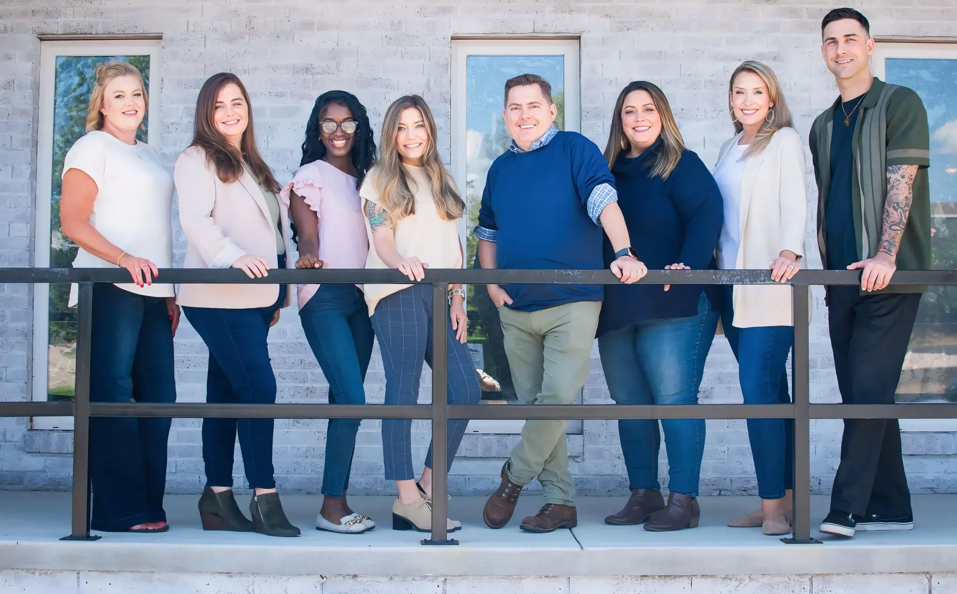 Eight people smile while leaning against a black railing in front of a light-colored brick building with windows.