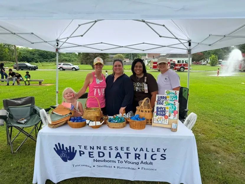 Volunteers at a Tennessee Valley Pediatrics booth under a white tent with baked goods and canned donations