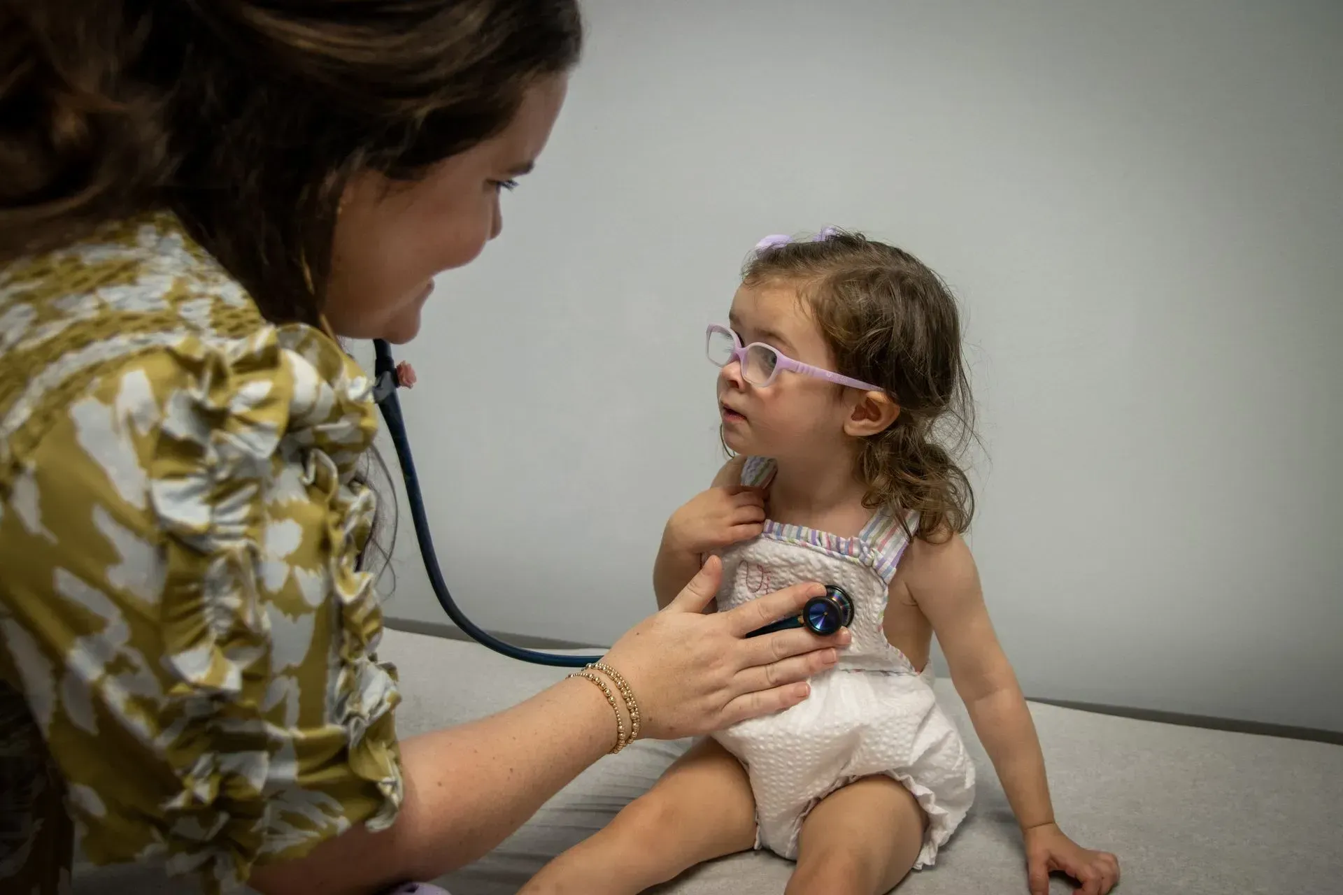 A healthcare provider uses a stethoscope to examine a young child wearing glasses in a clinic setting.