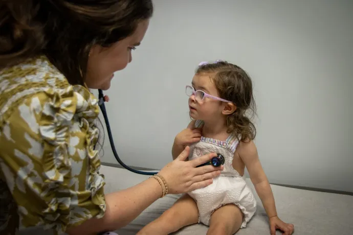 A healthcare provider uses a stethoscope to examine a young child wearing glasses in a clinic setting.