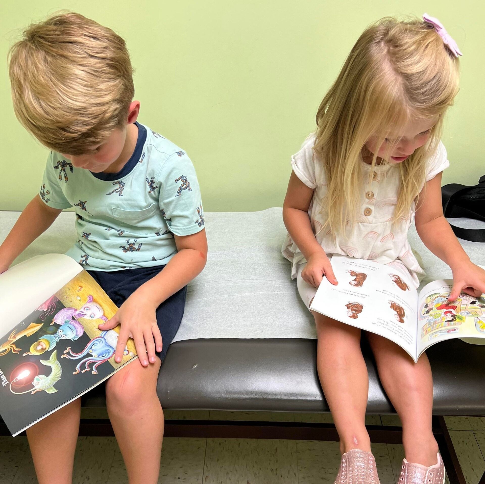 Two children sit side-by-side on an exam table, intently reading separate books in a doctor's office.