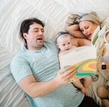 A parent reads a colorful book to a young child while lying together on a white bed.
