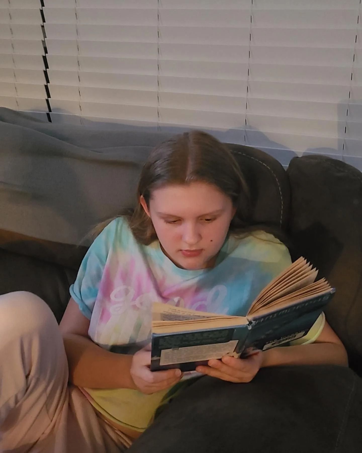 A person sits on a dark sofa, wearing a tie-dye shirt and reading an open book.