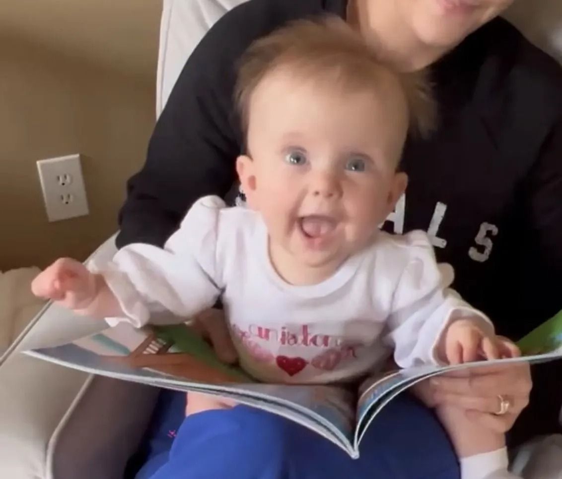 A smiling baby sits on a lap, holding an open book while facing the camera.