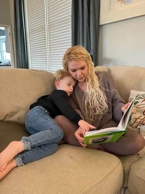 A person with long, blonde, wavy hair reads a book to a child while sitting together on a beige couch.