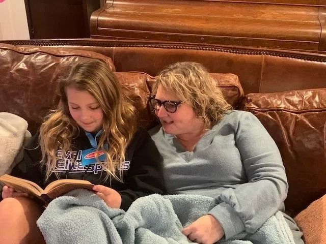A young person and an adult sitting together on a leather couch, reading a book while wrapped in a blue blanket.