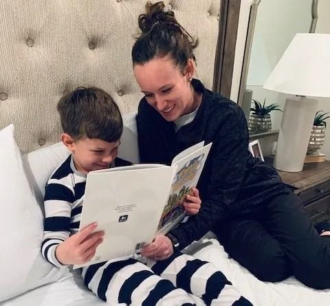 A person in dark loungewear sits on a bed reading a book with a smiling child in striped pajamas.
