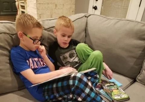 Two children sitting on a gray couch reading books together.