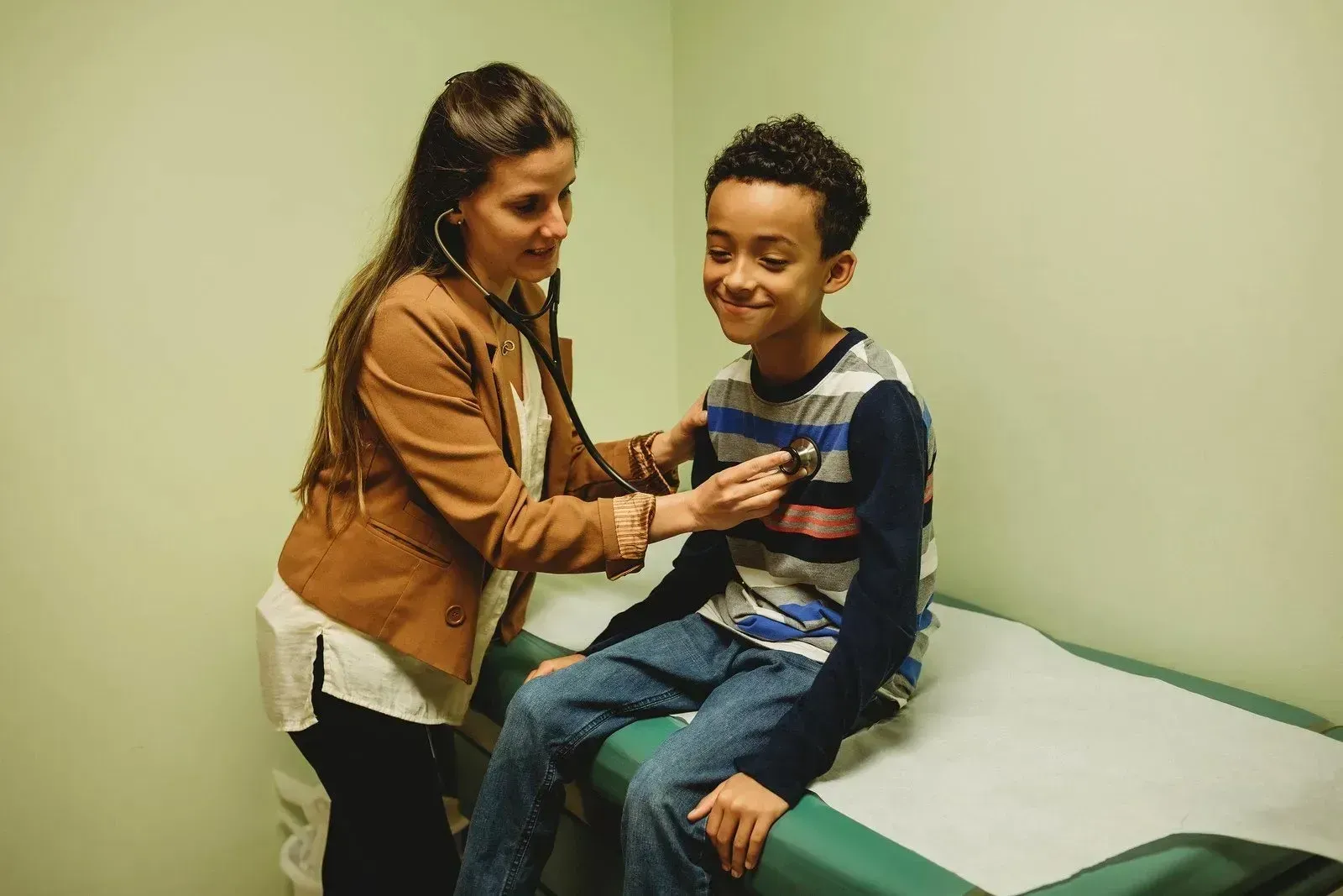 A healthcare provider in a tan jacket uses a stethoscope to examine the chest of a smiling child on an exam table.
