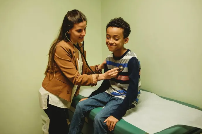 A healthcare provider in a tan jacket uses a stethoscope to examine the chest of a smiling child on an exam table.