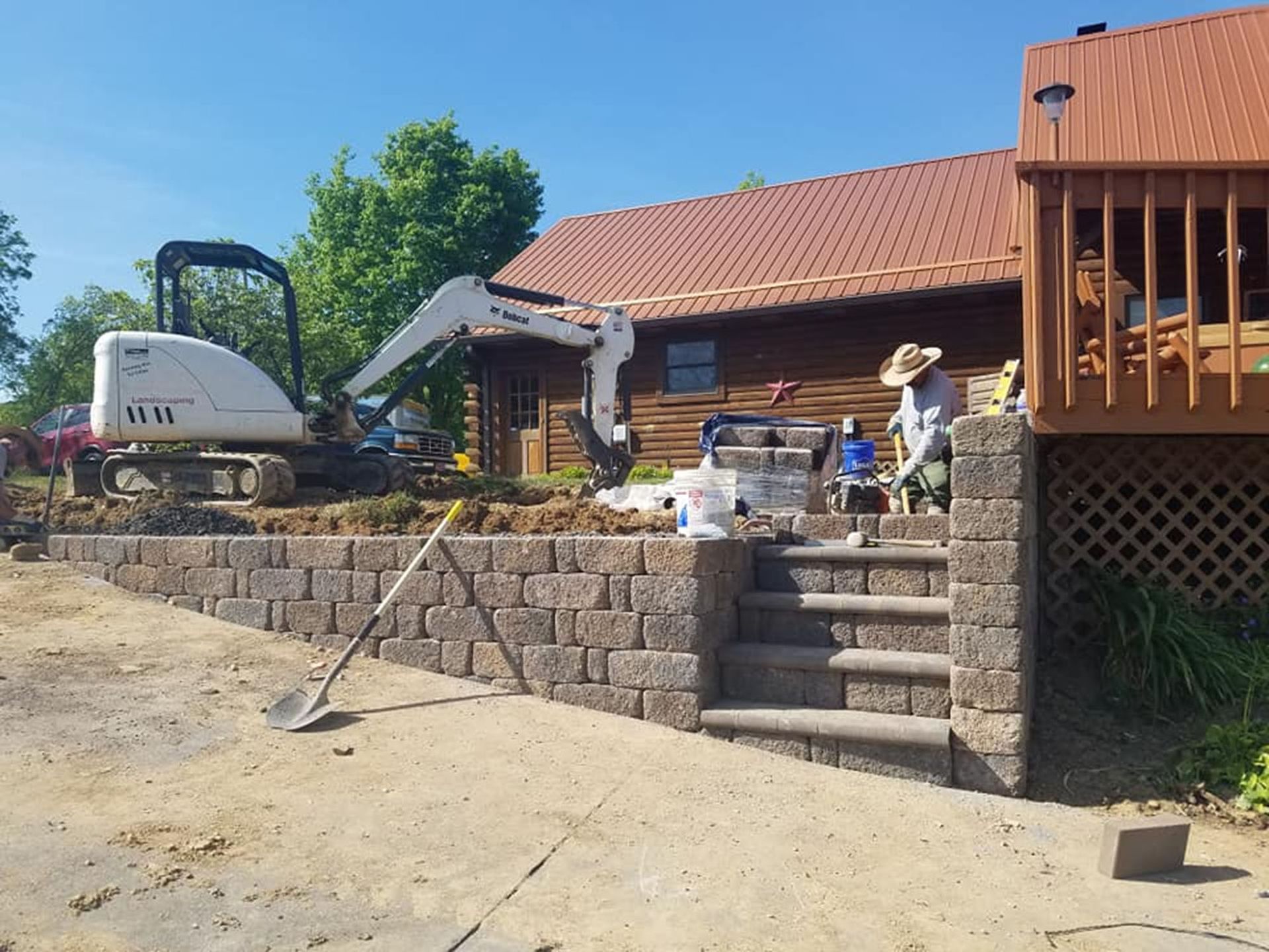 Stone retaining wall and steps leading to a log house with a red roof in a sunny yard under construction.