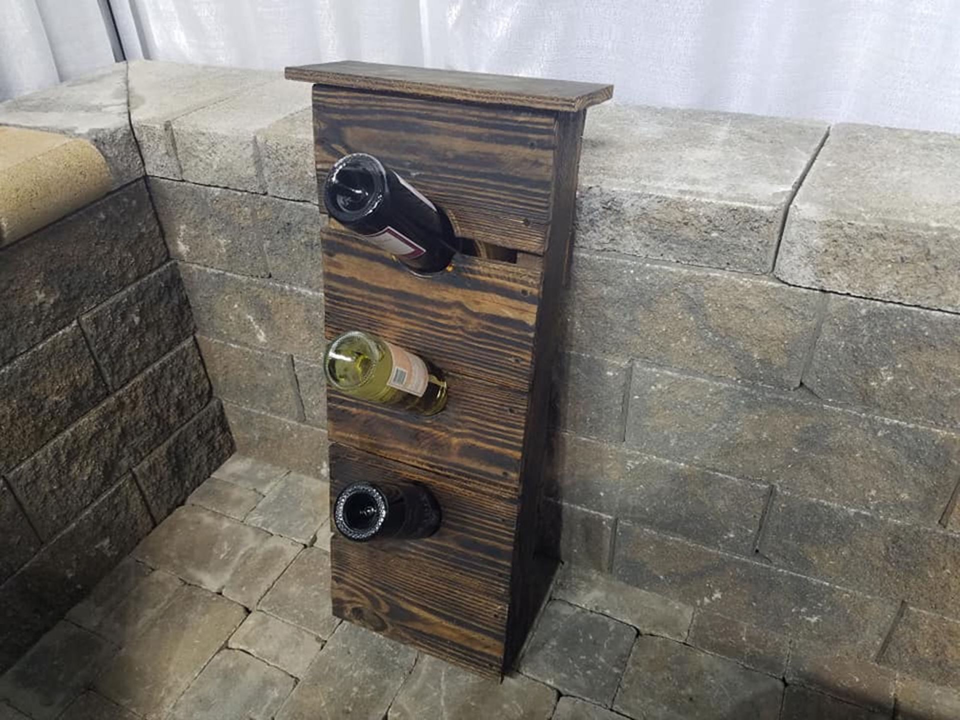 Three-bottle wine rack on a stone patio corner, holding two dark bottles and one white bottle.