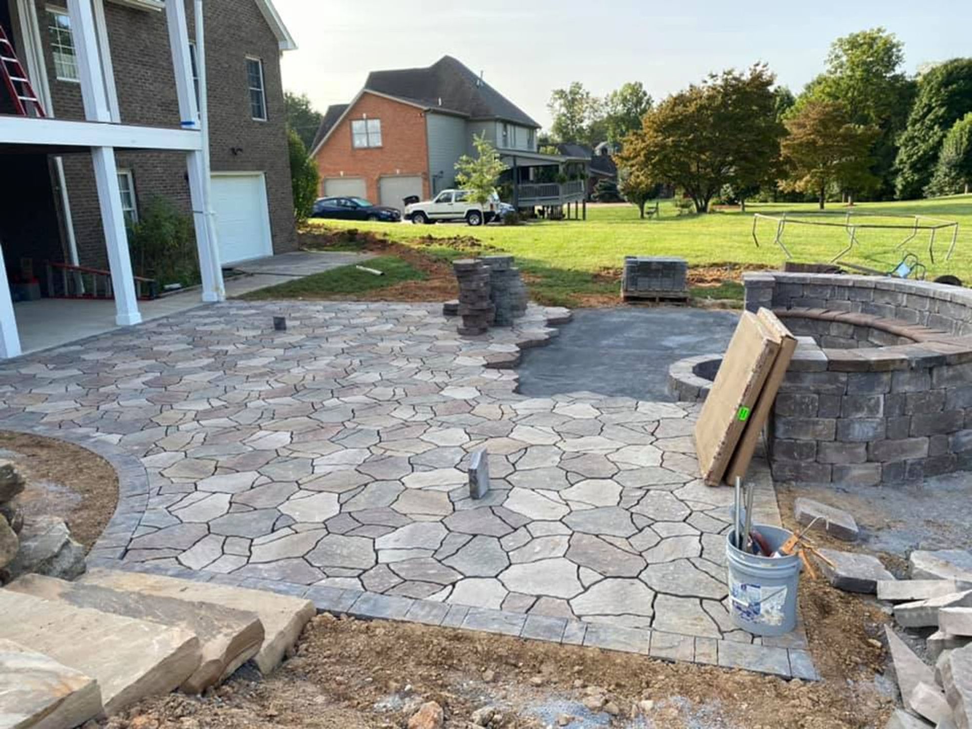 Backyard stone patio under construction with stacked pavers, buckets, and retaining wall blocks