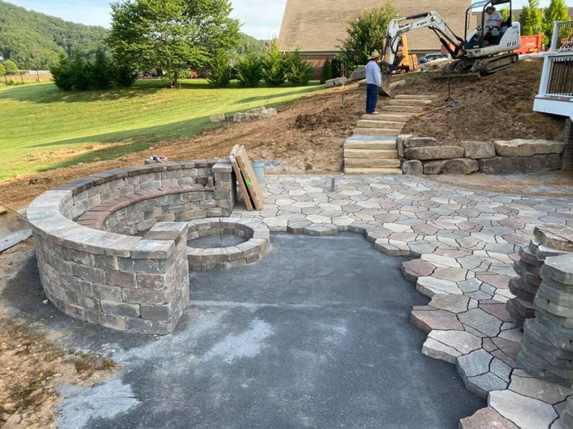 Stone patio and curved retaining wall under construction in a backyard, with workers and excavator nearby
