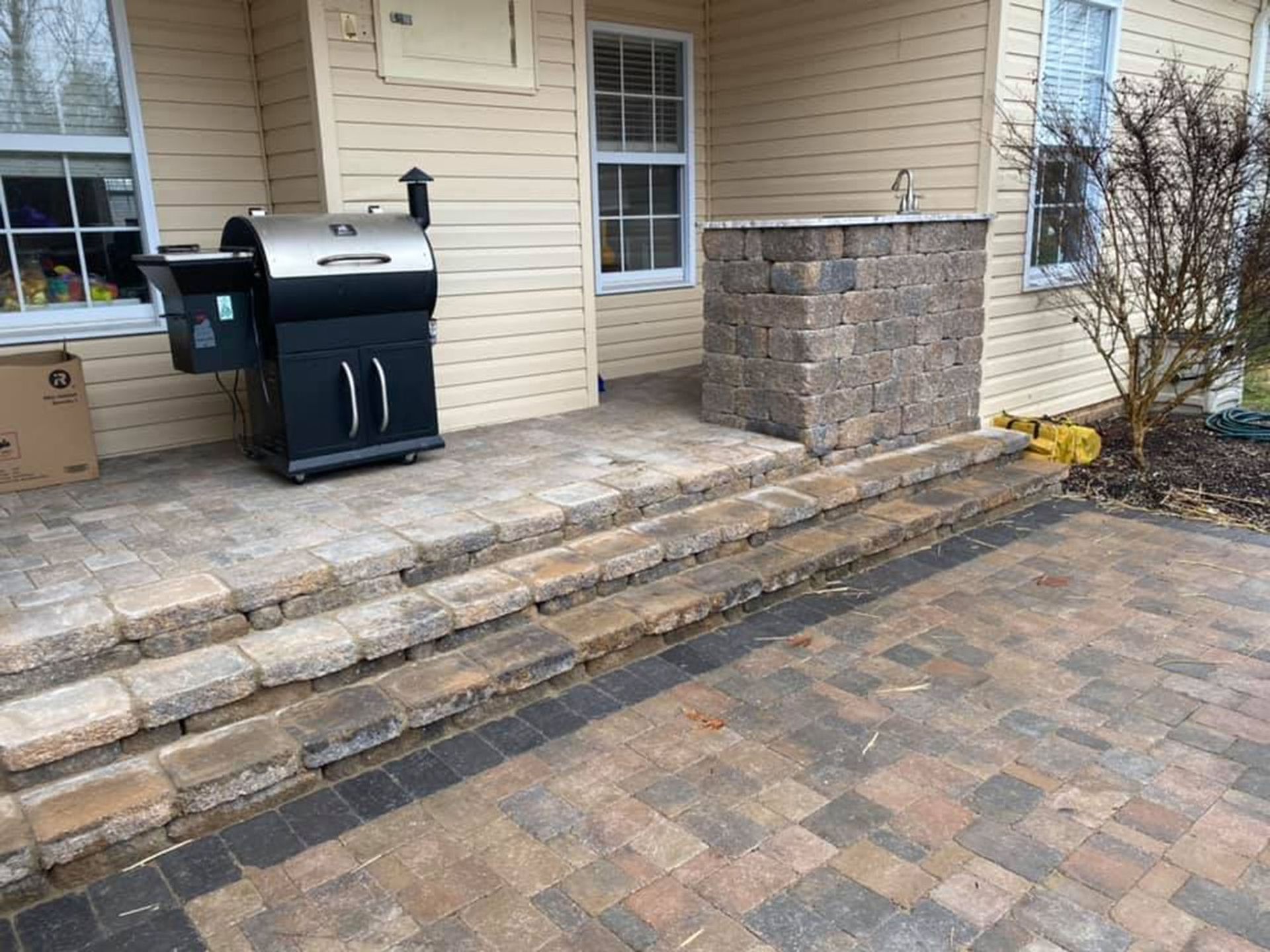 Patio with stone steps, brick pavers, a black grill, and a covered brick structure beside a house