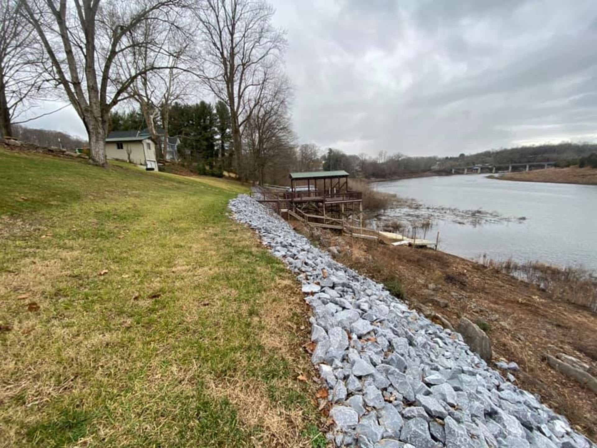 Rocky shoreline with a grassy bank, leafless trees, and a river under a cloudy sky