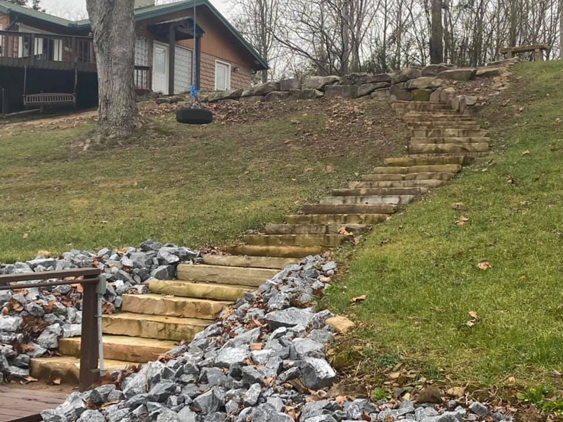 Rock steps bordered by piled stones lead up a grassy slope to a house in a wooded yard