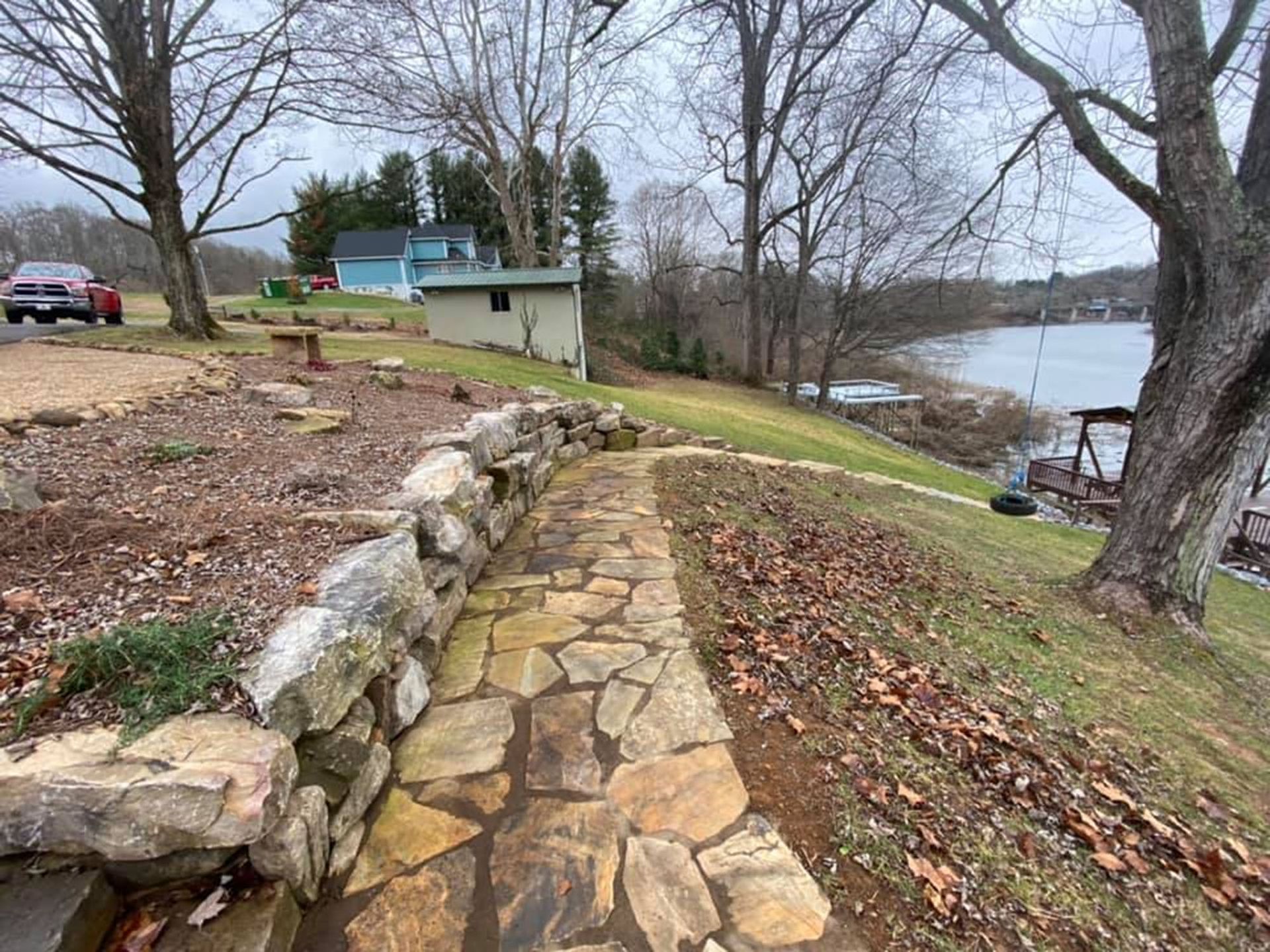 Stone path beside a lake with leafless trees and a small green cabin on a cloudy day