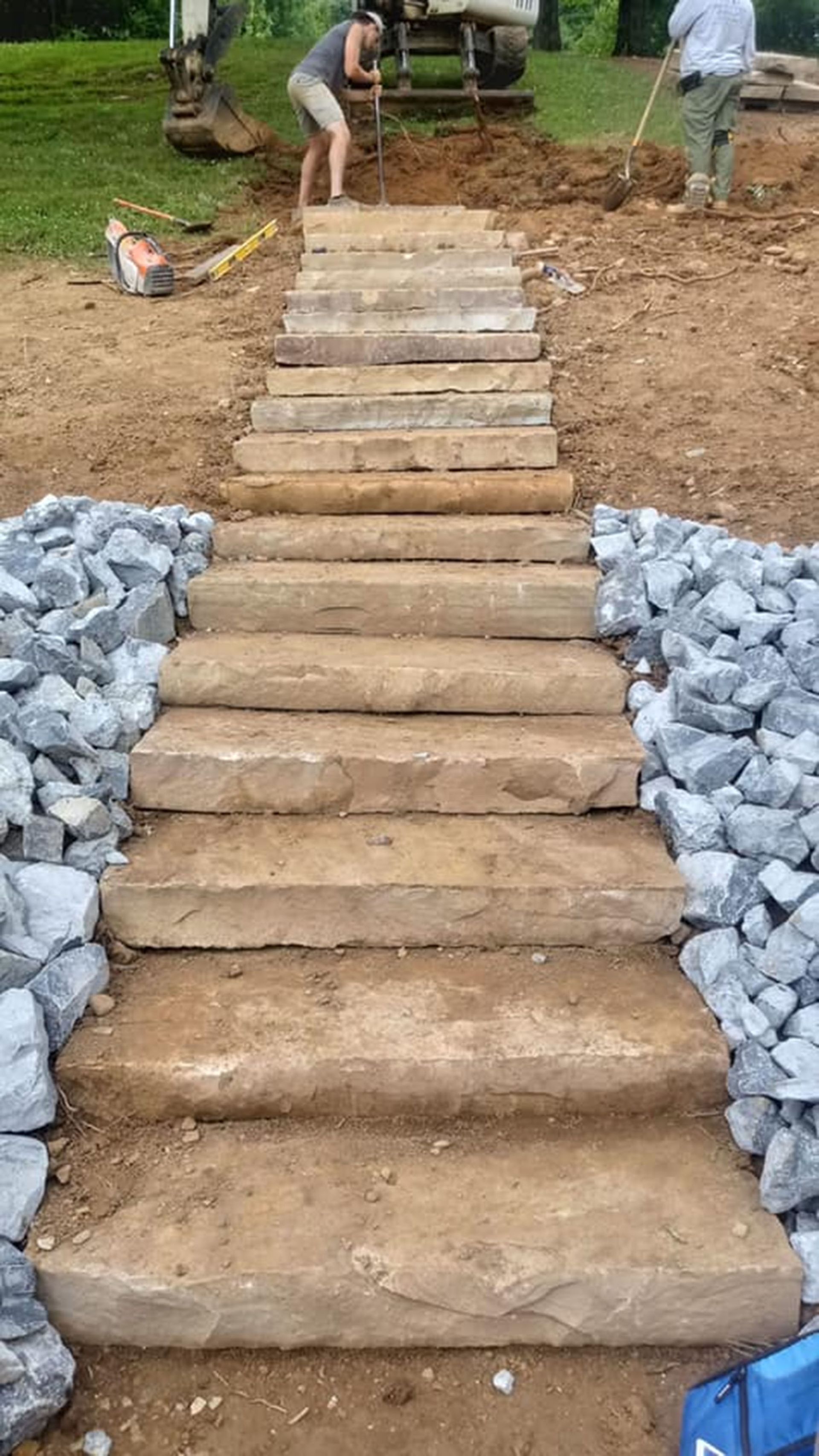 Stone steps under construction on a dirt path, with workers placing rocks and wood along the sides.