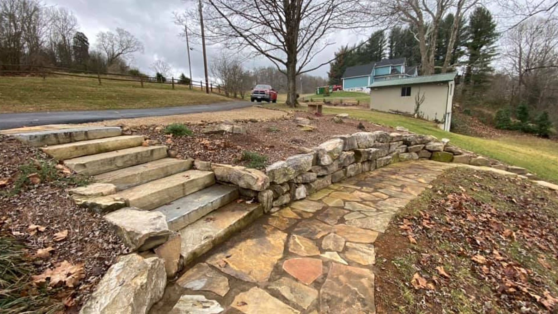Stone steps and a curved flagstone path in a yard, leading past a rock retaining wall to a small house.