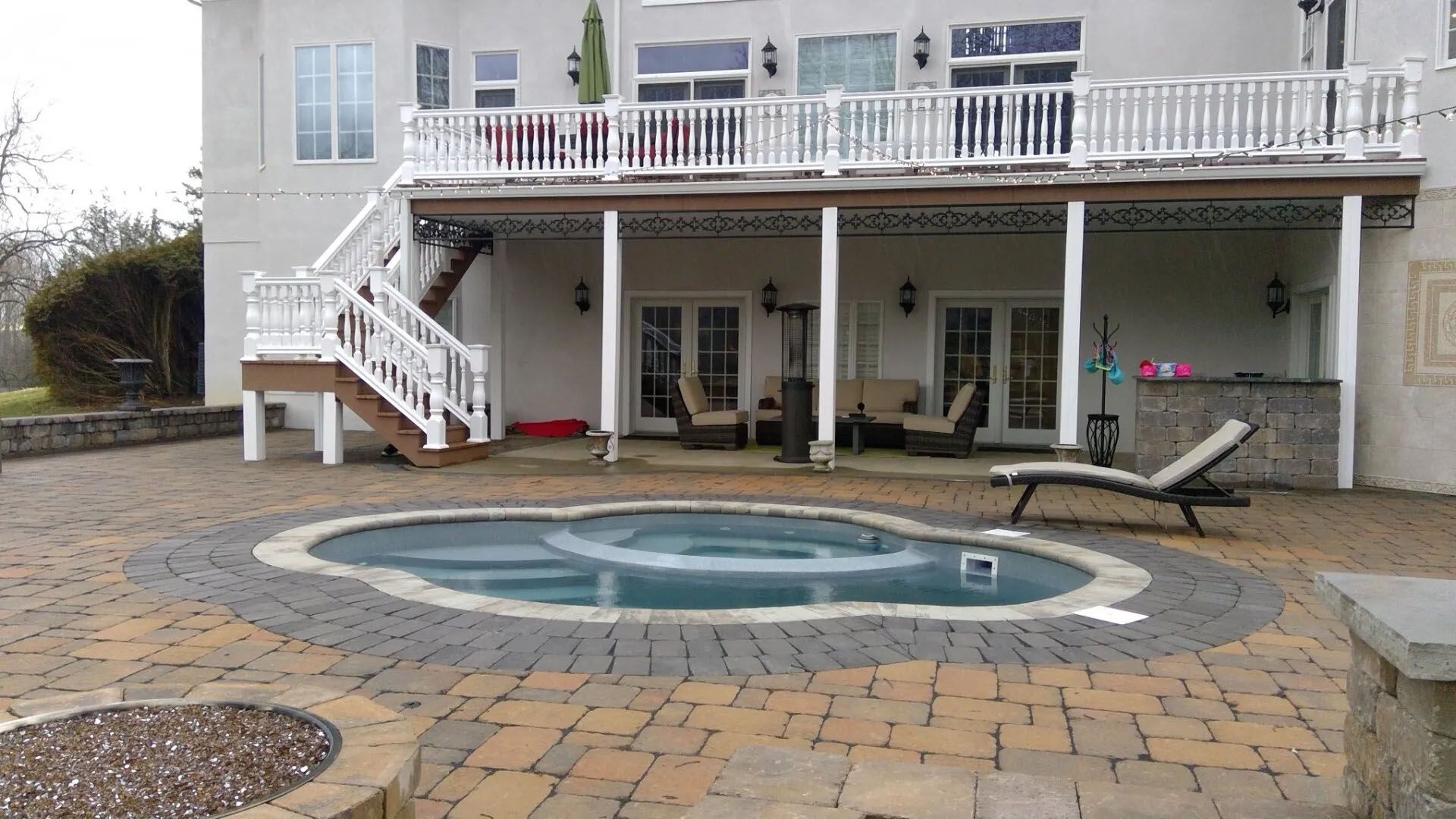 Backyard patio with a small round pool, stone pavers, and a raised deck with stairs and lounge chairs.