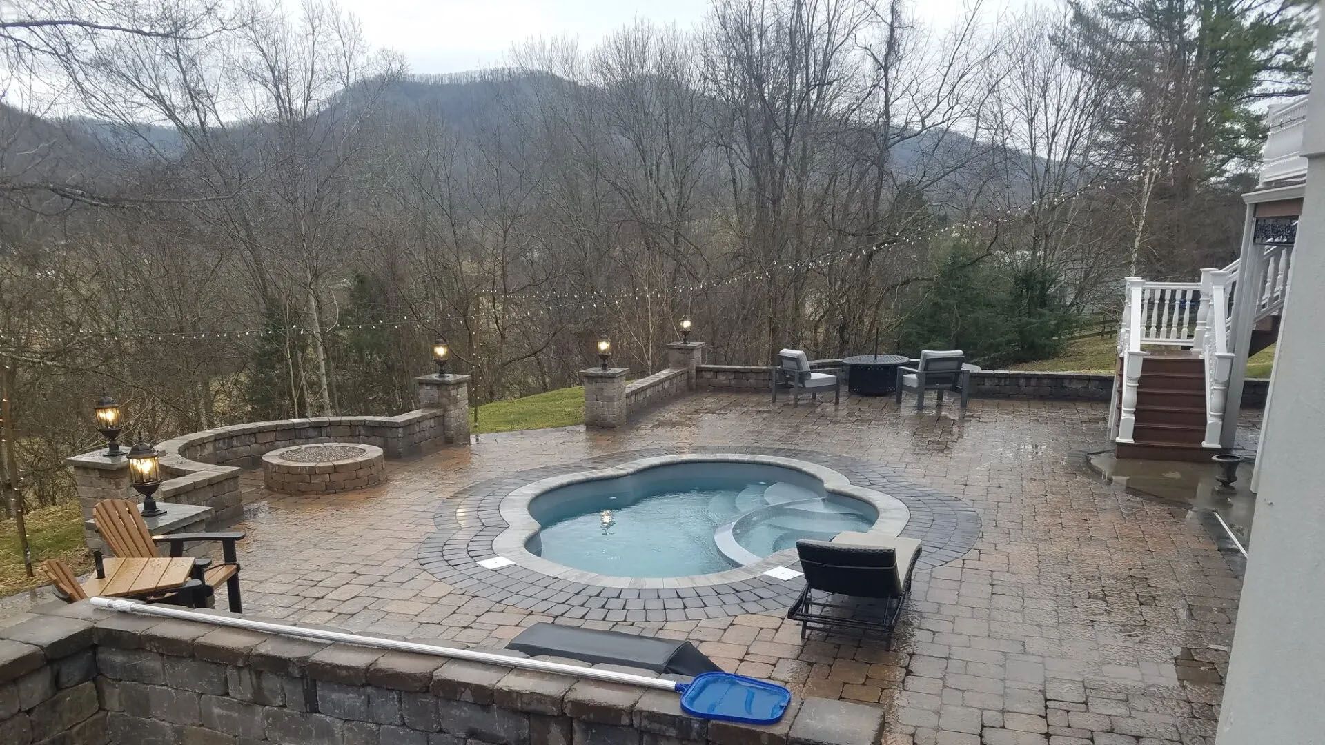 Rainy backyard patio with a small hot tub, stone seating, and wooded hills in the background