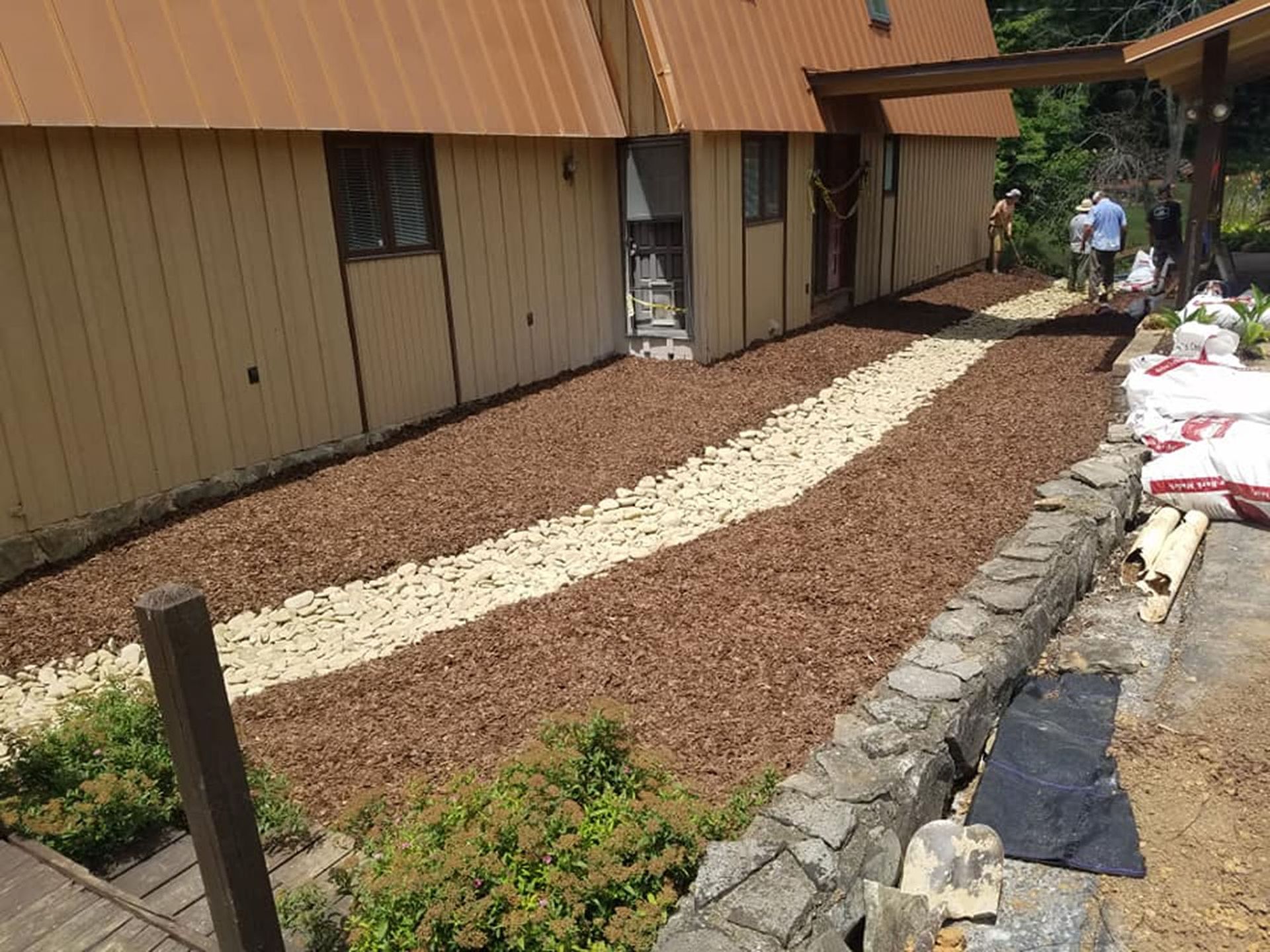 Garden path of white stones through brown mulch beside a building, with people and supplies in the background