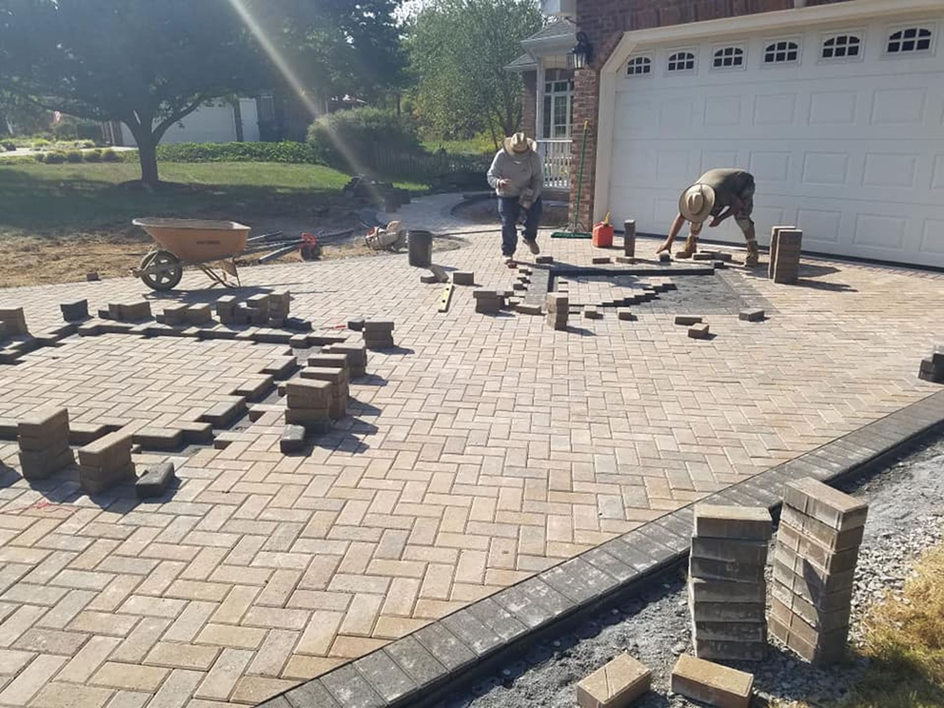 Workers laying pavers in a driveway beside a garage, with bricks and tools scattered around.