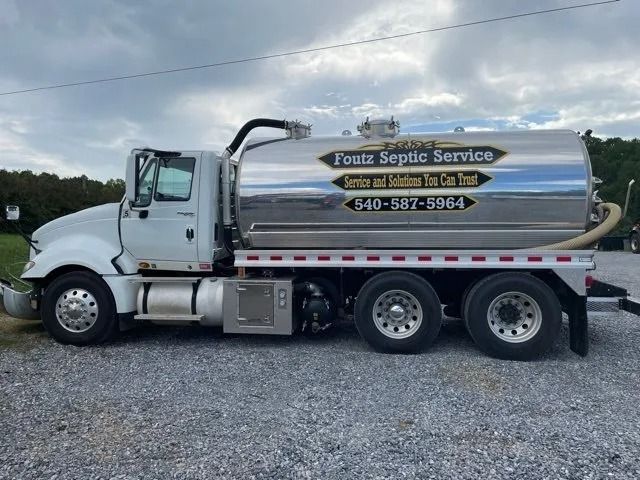 Silver utility tanker truck with company lettering parked on gravel under a cloudy sky