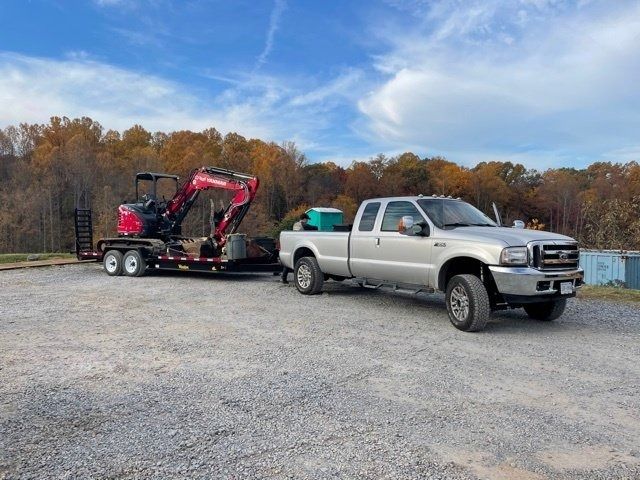 Silver pickup truck towing a red mini excavator on a trailer in a gravel lot under a blue sky