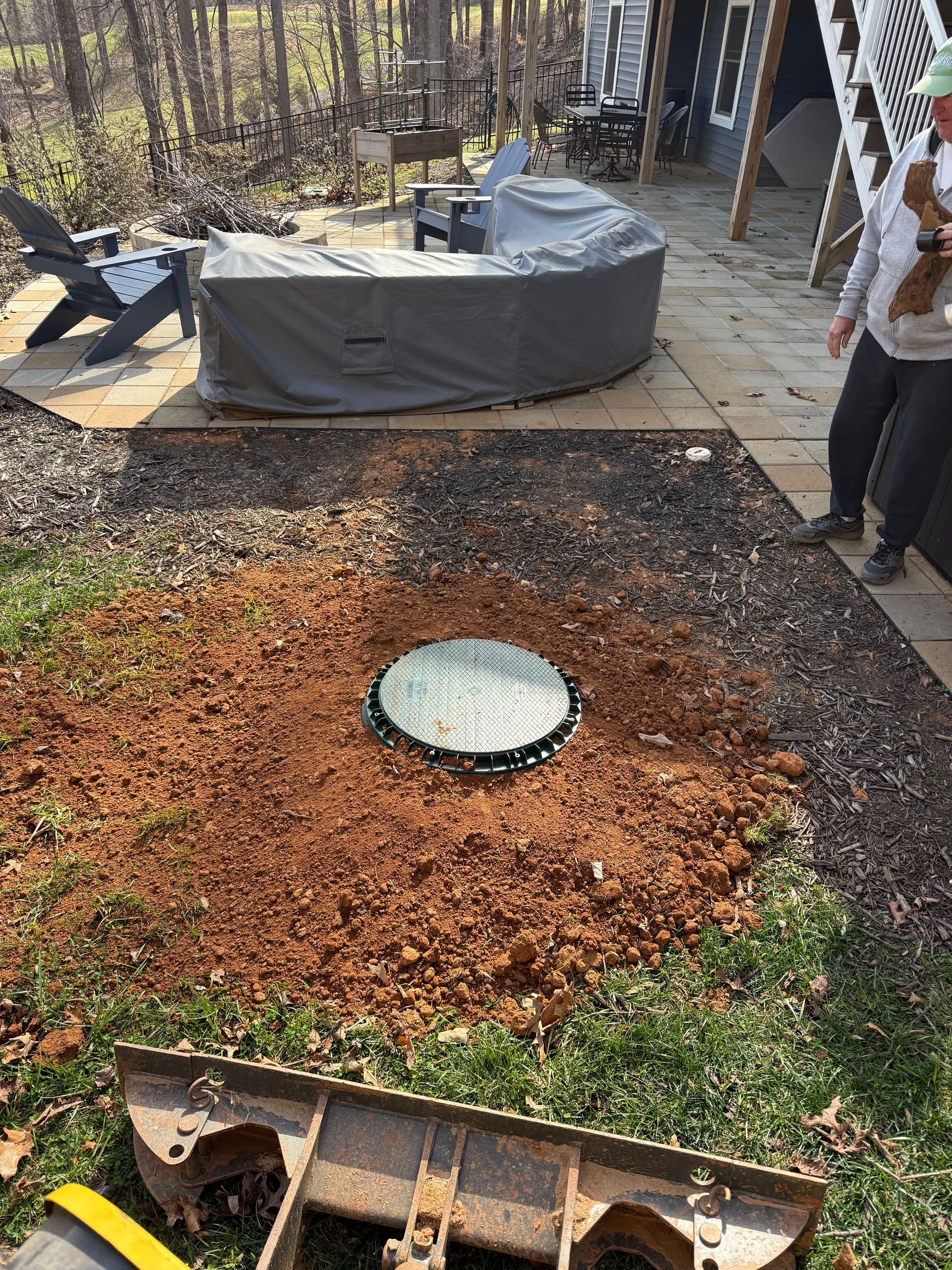 Backyard with circular mulch bed, round drain cover, covered hot tub, and a person standing at right