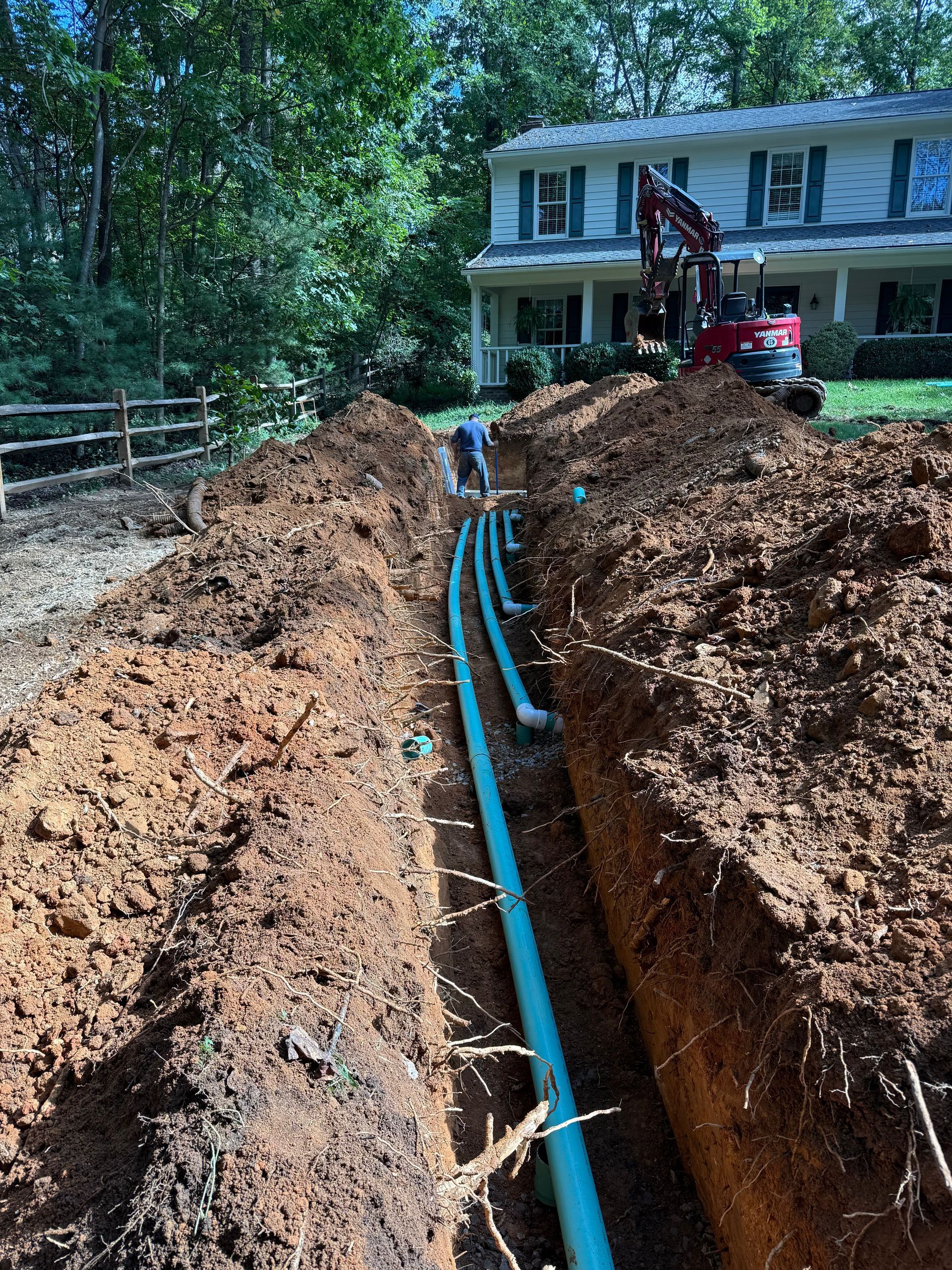 Trench with blue pipes being installed in a wooded yard beside a white house and excavator