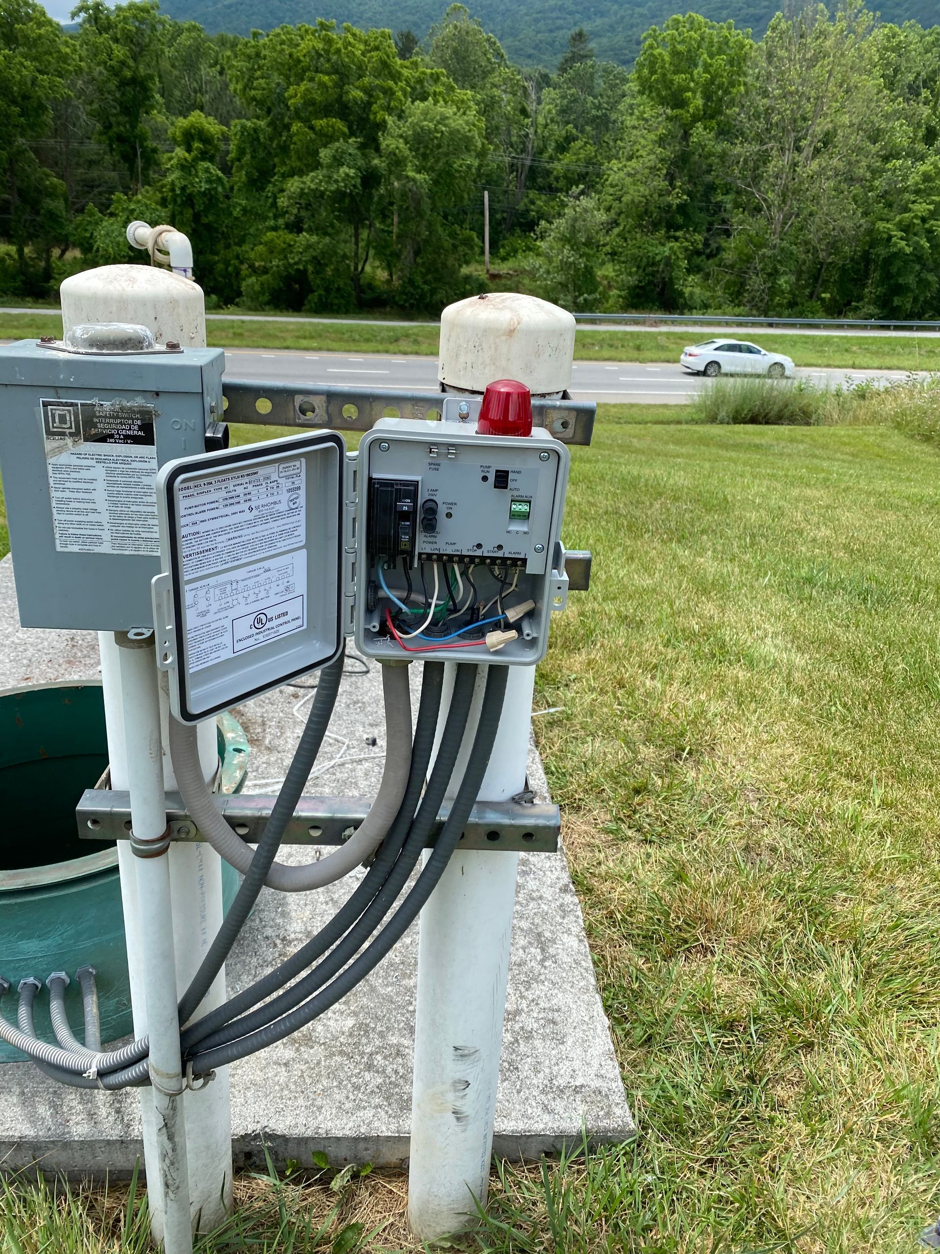 Open electrical control boxes and cables beside a grassy roadside under a wooded hillside