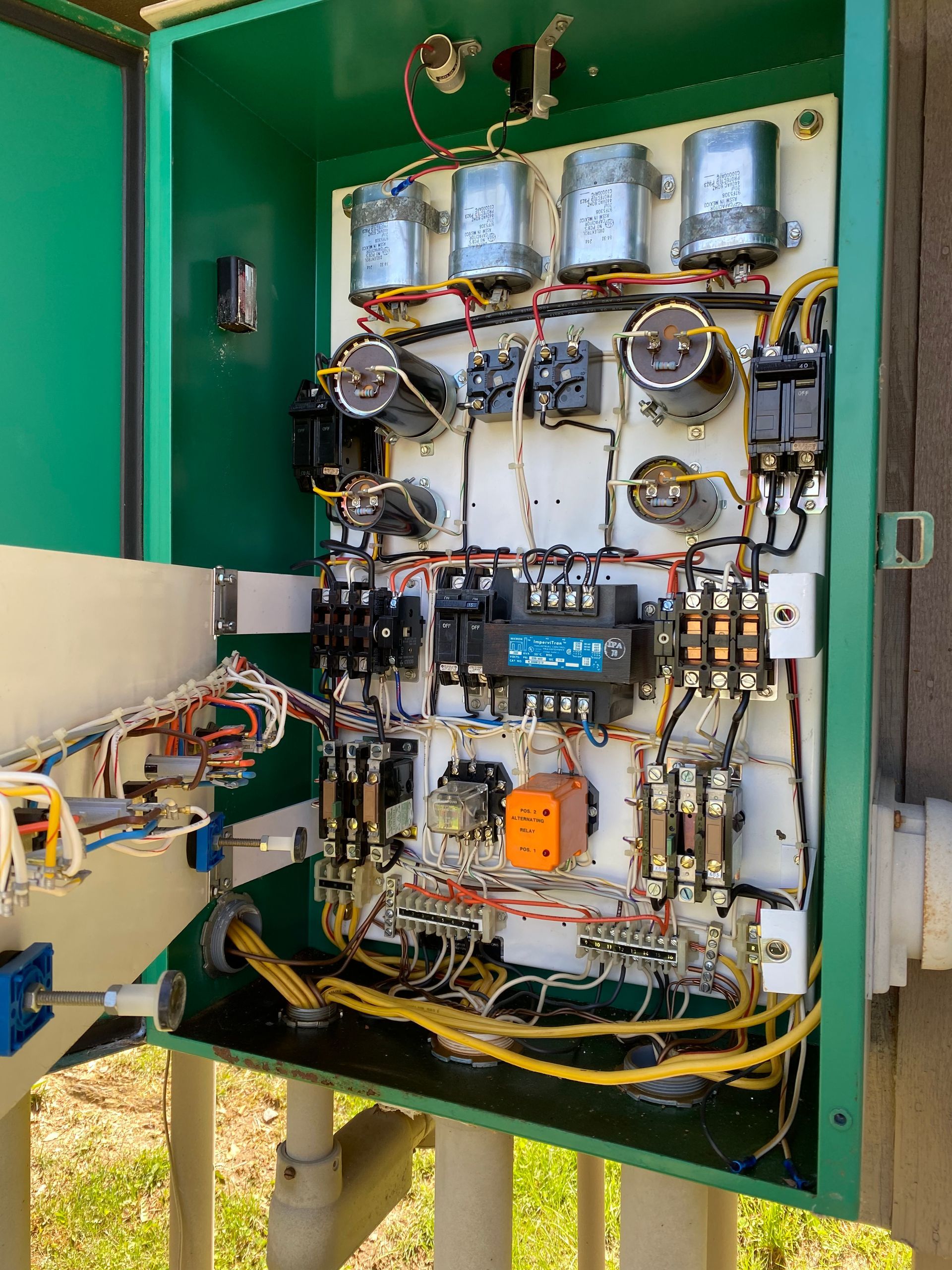 Open electrical control panel with wiring, relays, and gauges inside a green metal cabinet outdoors