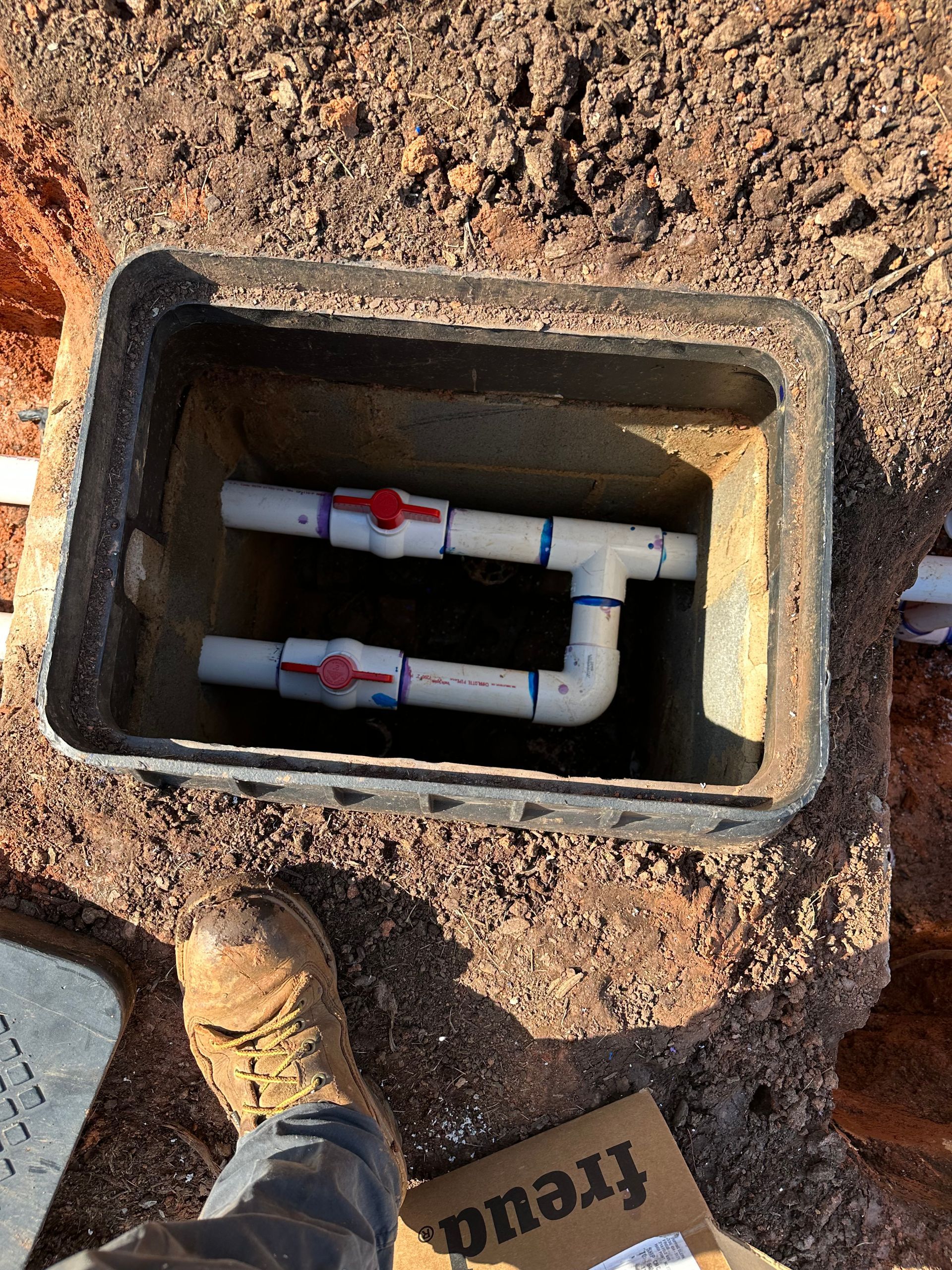 Open utility box with white pipes and red valves in dirt, beside a boot and a cardboard sign.