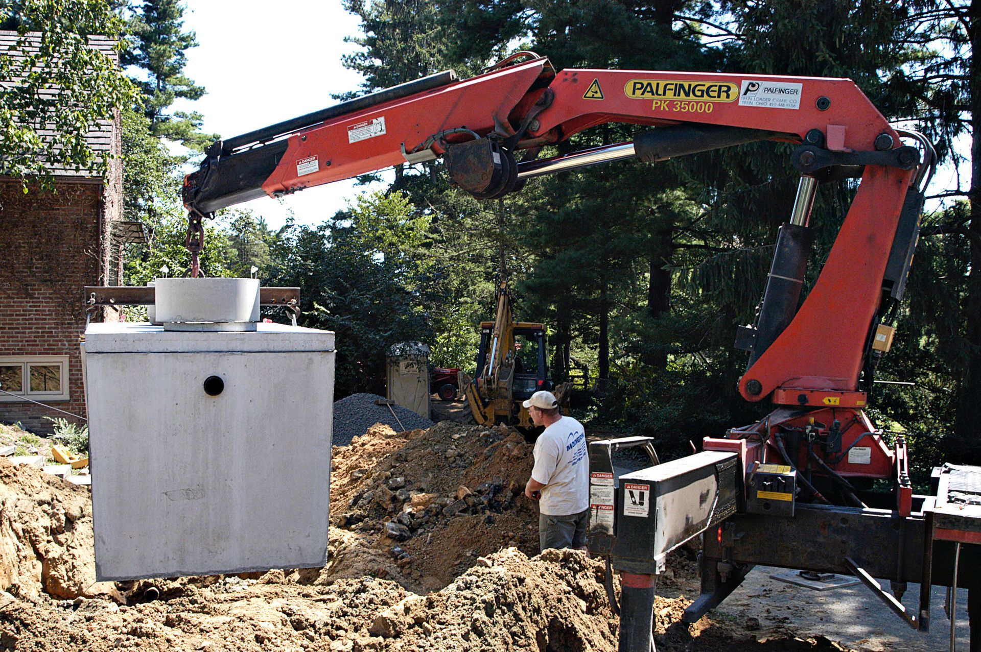 Red excavator lifting a concrete tank into a dirt excavation beside a worker in a wooded area