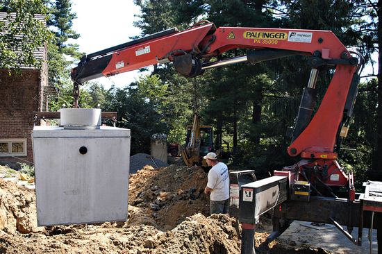 Red excavator lifting a concrete tank into a dirt excavation beside a worker in a wooded area
