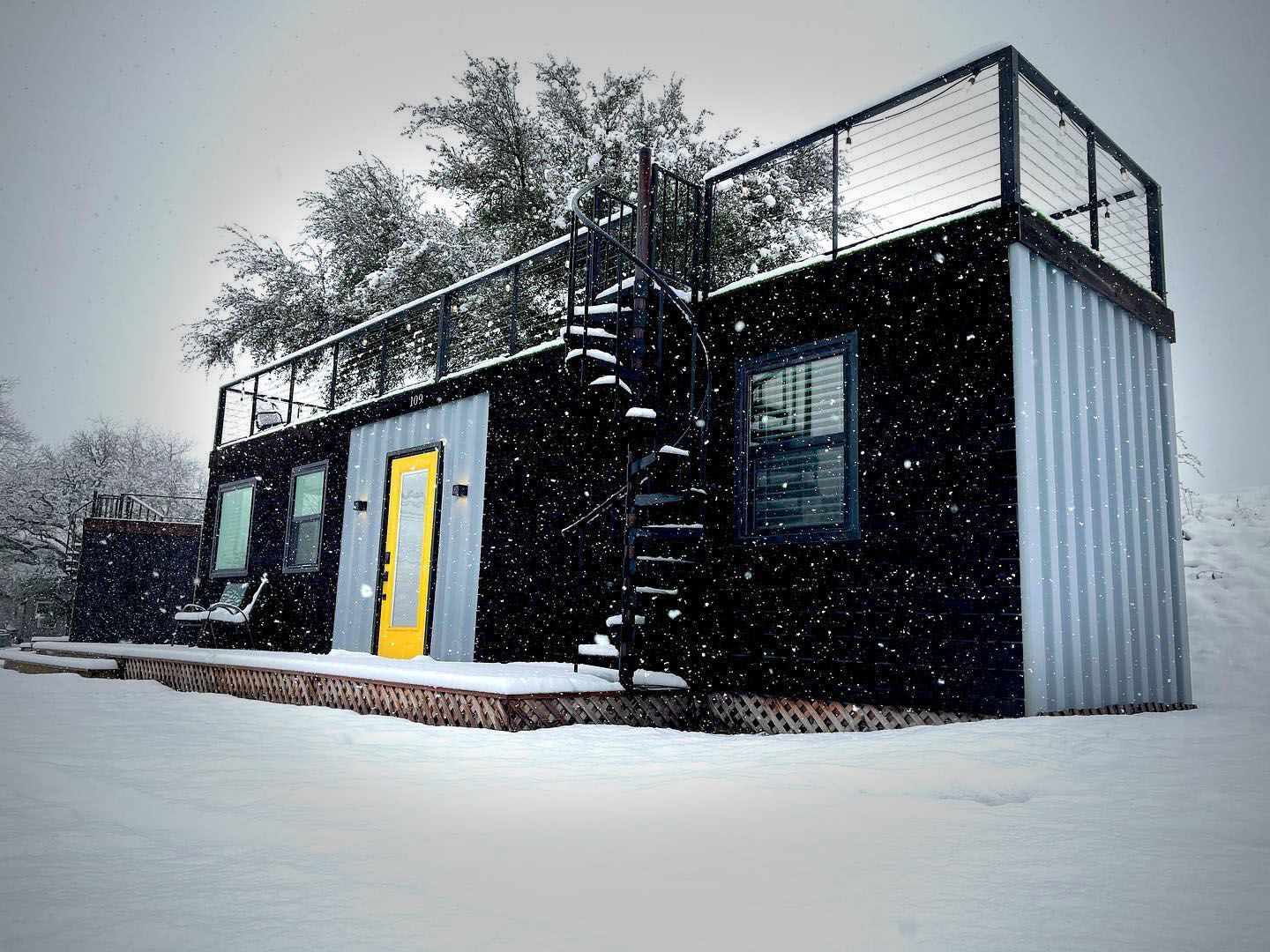 A small black house with a yellow door and stairs in the snow.