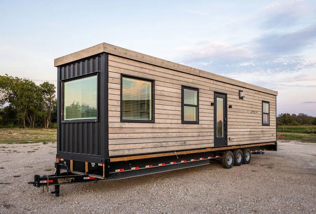 A tiny home on a trailer with wood siding and black trim, set on a gravel lot under a cloudy sky.