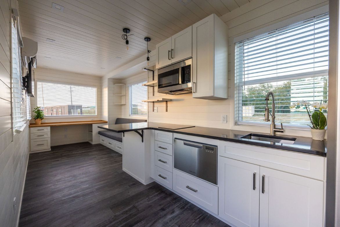 Interior of a tiny home kitchen with white cabinets, dark countertops, and a window with blinds.