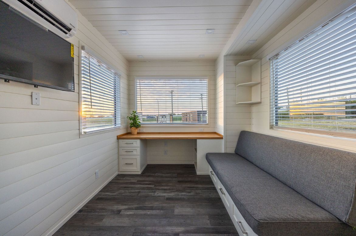 Interior of a small room with a desk, couch, and windows with blinds; white walls.