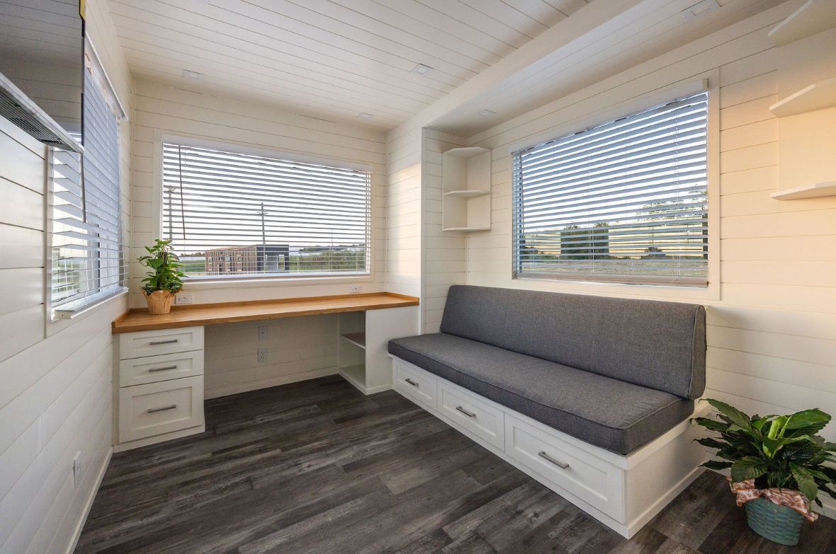 A small, bright room with a built-in desk, bench, and two windows with blinds. A plant is in the corner.