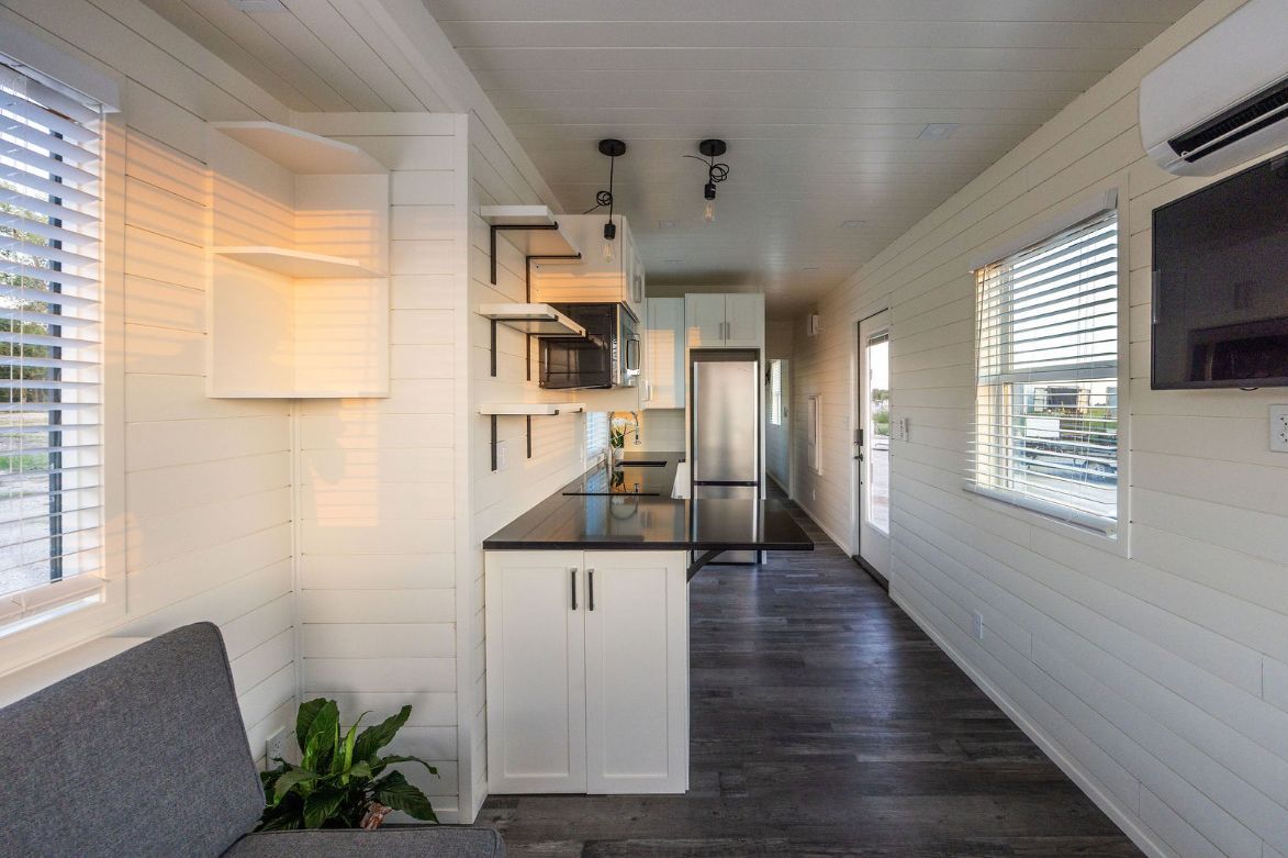 Interior of a tiny home with a kitchen, living area, and white walls. Dark wood floors and a gray sofa are visible.