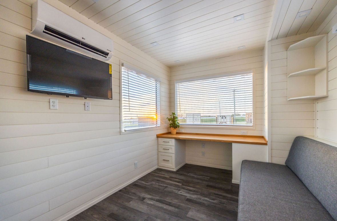 Small room with a desk, couch, window, and mounted TV. White walls and ceiling, wood-look floor.