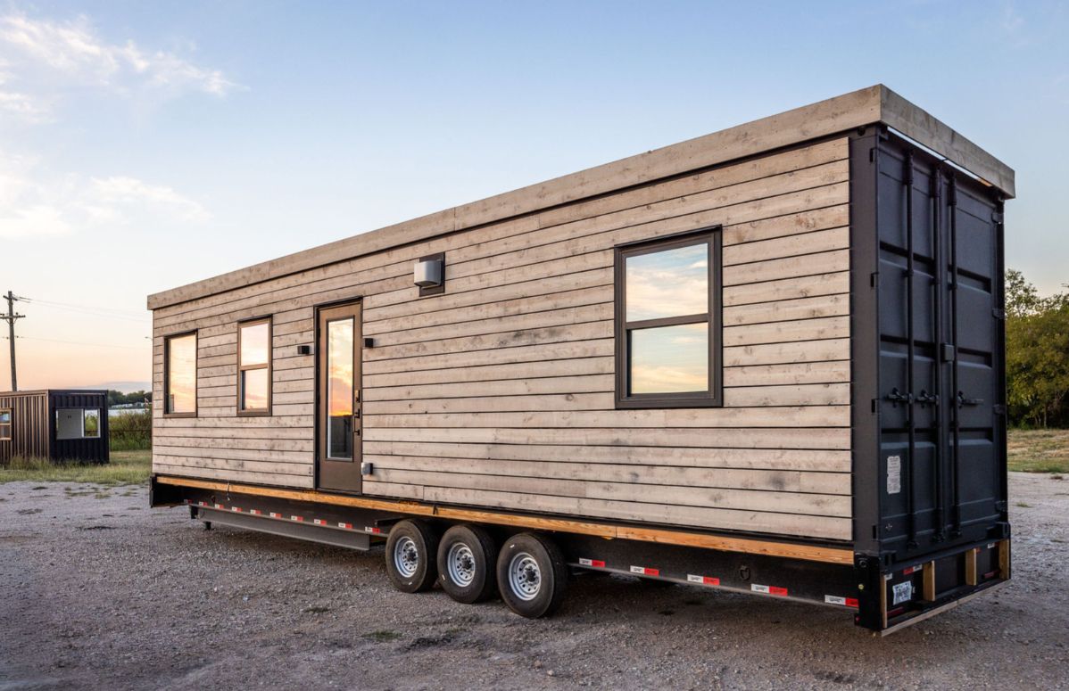 Shipping container home on a trailer, gray wood siding, black windows, three axles.