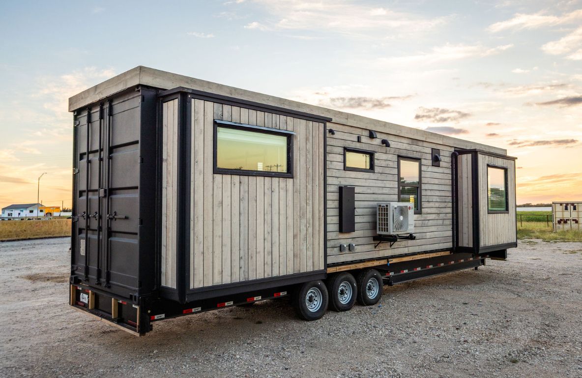 Modern tiny home on wheels with black and light wood siding, three axles, and windows.