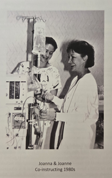Two women, Joanna and Joanne, in 1980s lab, examining a machine. One in a lab coat, smiling.