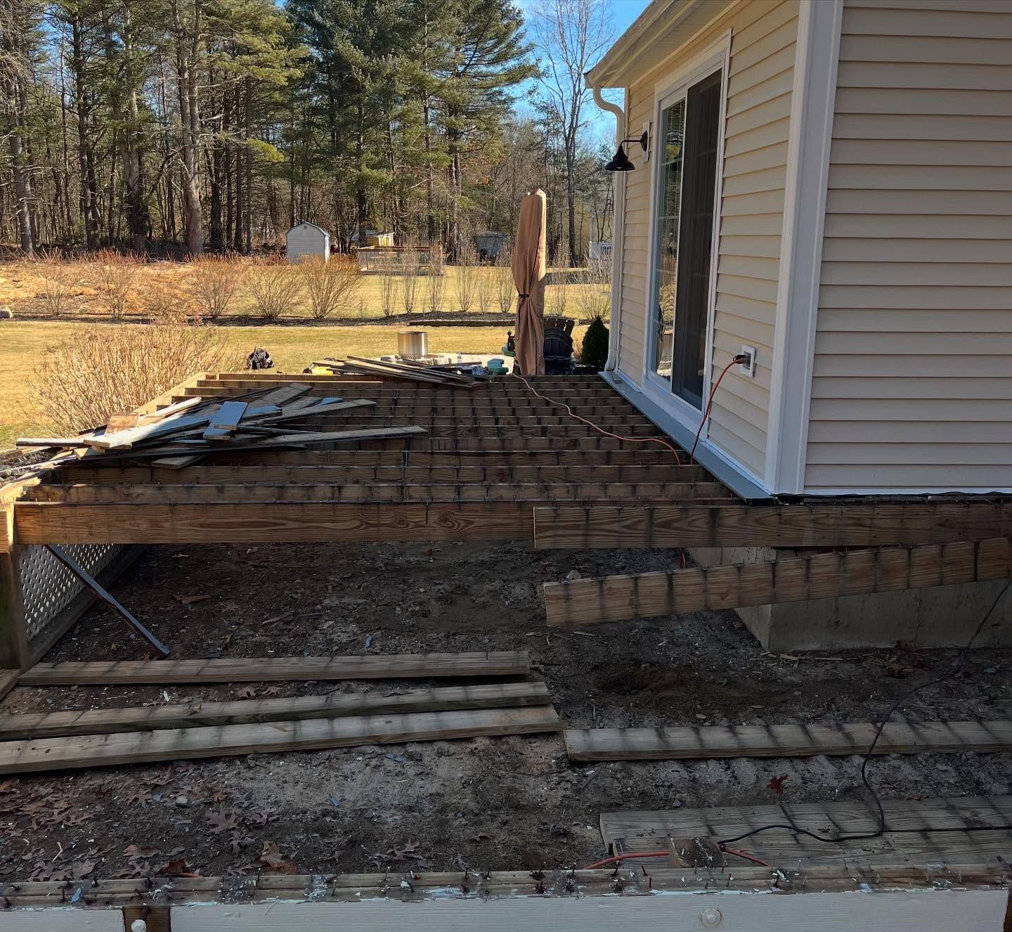 A side view of a house deck under construction with exposed wooden joists, set against a yard with trees and a shed.